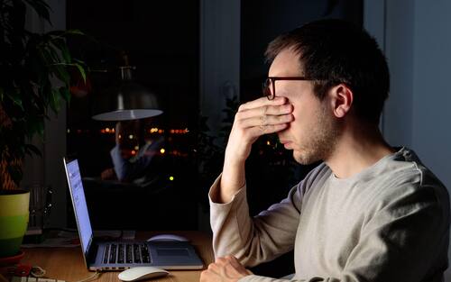 Tired freelancer man rubbing his eyes, sitting at desktop PC/laptop late at night, during the period of self-isolation and remote work at home, falls