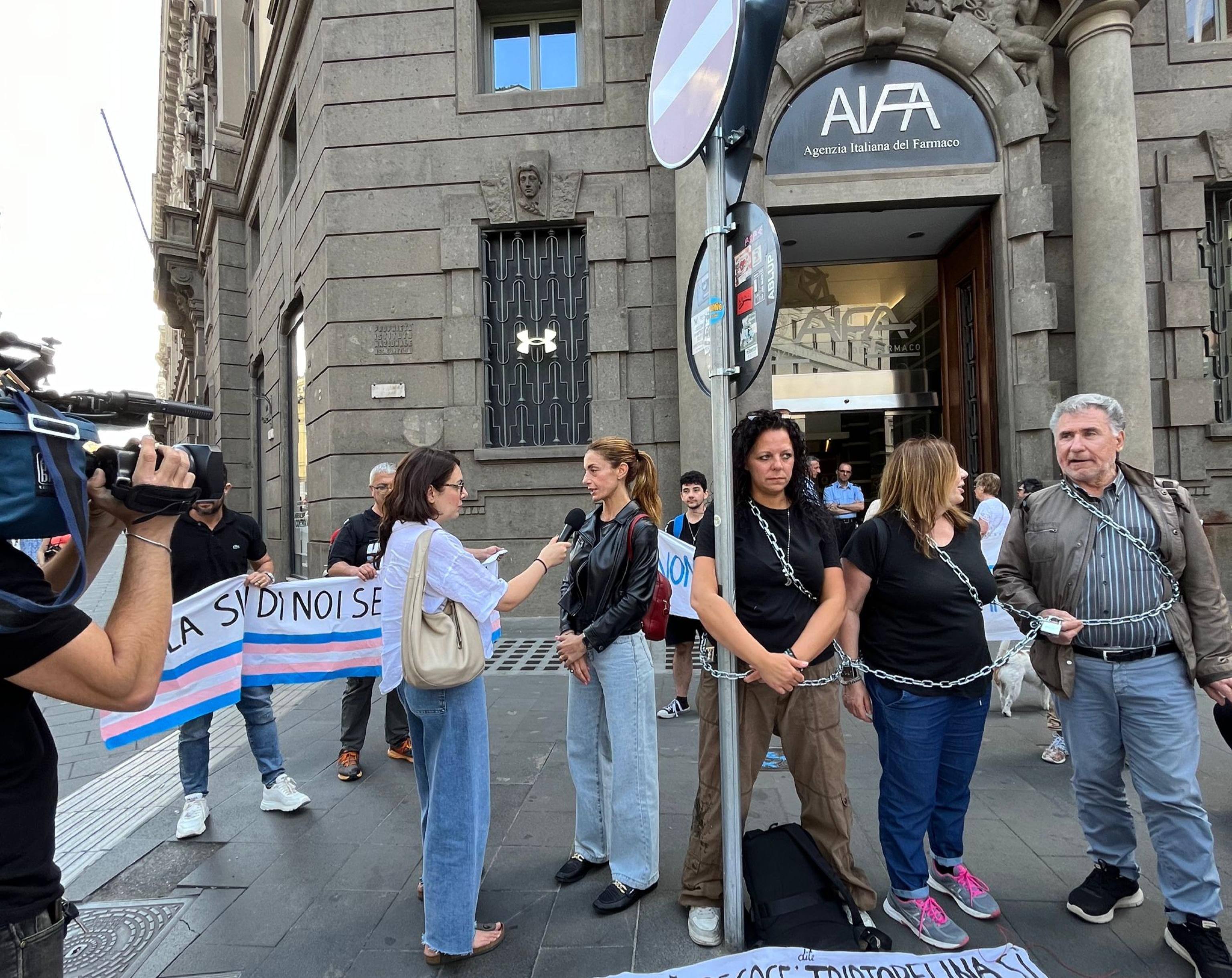 Una decina di persone, tra cui sei genitori con i figli adolescenti al seguito si sono incatenati simbolicamente oggi sotto la sede dell'Agenzia Italiana del Farmaco, a Roma per manifestare con contro il tavolo tecnico sulla prescrizione della Triptorelina, farmaco che sospende la pubertà, e utilizzato oggi per adolescenti con diagnosi di disforia di genere. Tra loro, a manifestare anche alcuni ragazzi stessi che indossavano la maglietta con la scritta: "Chiedimi se sono felice". "La vera domanda da porre, che però i politici che parlano di questo tema non si pongono, è proprio quella. Oggi siamo qui incatenati - lo dichiara all'ANSA Christian Cristalli, responsabile delle politiche trans nella segreteria nazionale di Arcigay - in occasione della prima riunione di un tavolo tecnico sui farmaci per la disforia di genere, perché a quel tavolo non ci sono associazioni, non sappiamo come siano stati scelti gli esperti. La triptorelina è un farmaco sicuro e dagli effetti reversibili, come conferma una nota stampa di 12 società scientifiche di pochi mesi fa. Ma su questo tema ora si fa campagna elettorale". "In pratica tutto viene fatto su di noi e senza di noi. Mentre siamo qui per dire che questo non è un tema su cui fare ideologia e propaganda, si tratta di salute e benessere delle persone", spiega all'ANSA Silvia mamma di Zoe, 14 anni e in trattamento da un anno, membro dell'associazione "Affetti oltre il Genere, creata da Cinzia Messina, una delle prime in Italia e ad esporsi sul tema. "In alcuni casi - aggiunge Silvia - si tratta addirittura di vita o di morte, perché ci sono adolescenti che per la difficoltà di questi percorsi sono arrivati a togliersi la vita mentre il farmaco li aiuta a gestire con più calma la gestione dei cambiamenti di un corpo in cui non si riconoscono".
ANSA/Associazione Affetti Oltre il Genere  ANSA PROVIDES ACCESS TO THIS HANDOUT PHOTO TO BE USED SOLELY TO ILLUSTRATE NEWS REPORTING OR COMMENTARY ON THE FACTS OR EVENTS DEPICTED IN