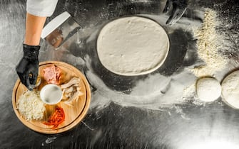 Cooking pizza on a steel table; dough, flour, pizza topping, sauce and chef`s hands in black gloves on the table