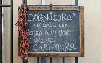 “Sognatore è colui che dentro a un uovo ci vede una carbonara (A dreamer is the one who sees a carbonara inside an egg)”, written on a black chalkboard with red chili peppers; sign outside a restaurant in Rome, Italy, Europe, European Union, EU. - Close-up.
Carbonara is a typical Italian pasta dish from Rome made with eggs, hard cheese, cured pork, black pepper.
