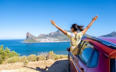 Woman outside car window road trip with hands up, car at Chapmans Peak Drive in Cape Town South Africa looking out over ocean