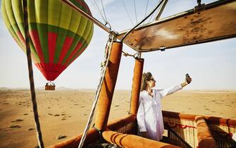 Wide shot of smiling mature woman taking selfie during early morning hot air balloon ride over the deserts of Morocco