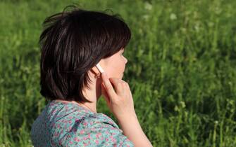 Woman touches wireless earphone in the ear standing on summer meadow. Headset, listening to music and voice call on a nature