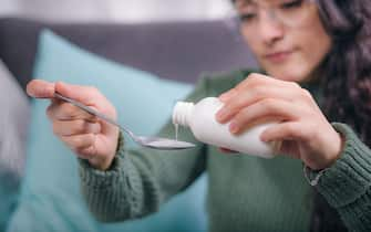 Medicine concept. Young woman's hands holding a cough syrup on a spoon, the woman is sitting on an armchair in her house.