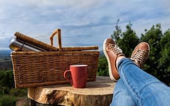 Woman relaxing in nature. Woman's feet, picnic basket and mug on nature background. Glamping.