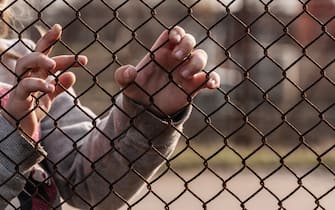 The child's hands are holding onto a metal mesh fence. Social problem of refugees and forced migrants.