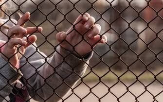 The child's hands are holding onto a metal mesh fence. Social problem of refugees and forced migrants.