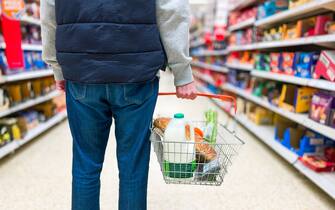 Low angle close up color image depicting a man holding a shopping basked filled with essential fresh groceries like bread and milk in the supermarket.