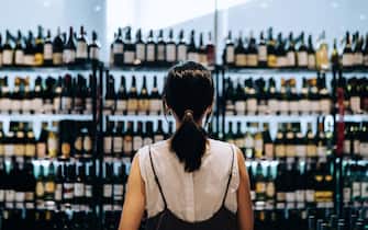 Rear view of young Asian woman grocery shopping for wines in a supermarket. She is standing in front of the liquor aisle and have no idea which wine to choose from