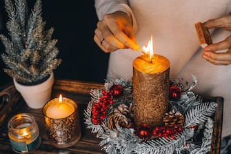 woman hands lightning big gold candle with matches in Christmas wreath. Xmas interior decoration on rustic wooden background. hygge, ornaments, candle