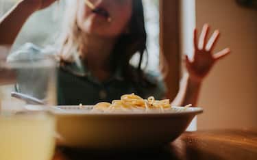 A bowl of spaghetti pasta on a table. A child is in soft focus in the background, slurping some spaghetti.