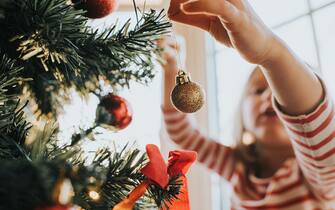Cute little girl reaches up and carefully places a gold glittery bauble on the Christmas Tree. Festive and warm image with space for copy.