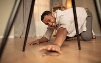 Handsome young black man standing on his knees, trying to reach house keys, dropping them on floor at home. Millennial guy being careless or forgetful, losing his things indoors