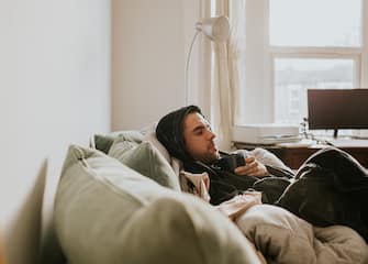 A man in a domestic living room relaxes on a comfortable sofa. He is wearing a dressing gown with the hood up, over his head. He holds a mug and enjoys a hot beverage.