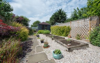 A view looking down a back garden of a home with paving slaps and gravel, pea shingle, wooden railway sleeper flower bed, vegetable patch, potted plants, timber fence and grey summerhouse, shed with tiled roof on a warm sunny day