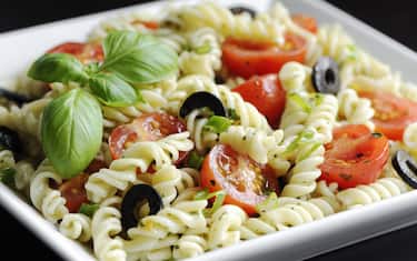 "Fusilli pasta salad with cherry tomatoes, chives, black olives, olive oil, basil leaves and spices in a white bowl."
