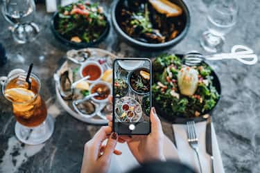 Overhead view of young woman taking photos of scrumptious and delicious meal on dining table with smartphone before eating it in restaurant. Eating out lifestyle. Camera eats first culture
