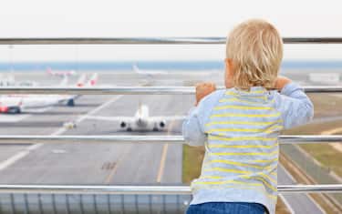 Little baby boy waiting for boarding to flight in airport transit hall and looking at airplane near departure gate
