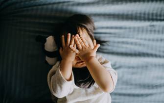 Portrait of cute little Asian toddler girl covering her face playing peekaboo while lying on bed with panda soft toy lying next to her