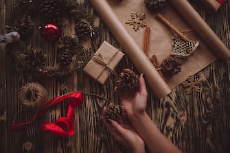 Top view of female hands holding wrapped gift box with ribbon on table surrounded by paper, christmas toys and garland.