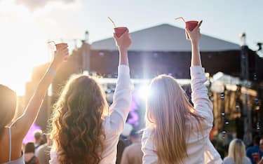 Party atmosphere with lively crowd, outdoor event. Group of girls at a beach music festival, holding drinks up, enjoying the sunset. Female friends celebrate, dance near stage, summer vibes.