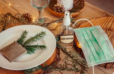 Christmas eve holiday party decorated table set with disposable medical mask and alcohol hand sanitizer bottle. Coronavirus (Covid 19) spreading prevention concept. Christmas micro led lights wire.