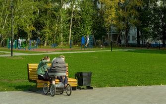 Two women talk on a park bench while rocking a newborn baby in a pram.