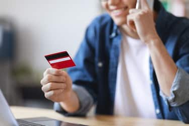 Mobile Payments. Smiling Young Male Holding Credit Card And Taling On Cellphone While Sitting At Desk In Office, Unrecognizable Millennial Man Enjoying E-Commerce And Easy Shopping, Cropped
