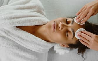 Professional beautician cleansing skin with cotton pads to dark-skinned young woman.
