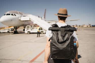white young man with hat and suitcase walking through the airport near the takeoff runway