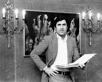 Frontal portrait of the Italian actor Lando Buzzanca wearing a pied de poule pattern jacket and pair of jeans, and standing in front of two wall lamps and a symbolist painting; the actor is holding a photo album. Rome (Italy), 1974. (Photo by Mario Notarangelo/Mondadori via Getty Images)