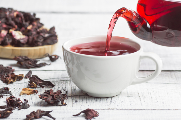 White cup of healthy hibiscus tea pouring from the teapot with dried hibiscus flowers on white wooden background, winter hot drink concept for cold and flu