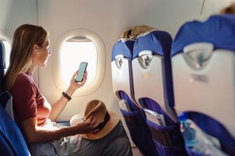Young woman sitting on the aircraft seat near the window during the flight in the airplane. She is using smart phone with a blank screen