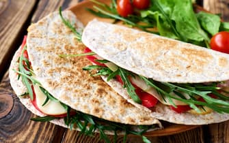 Piadina Romagnola with mozzarella cheese, tomatoes, ham and rocket salad on wooden table. Italian flatbread or open sandwich. Selective focus.