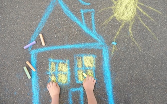 Little child boy is drawing house and sun painted with colored chalk on asphalt of sidewalk. Kids creative picture on gray background of road. Concepts of home and peaceful life all over the world.