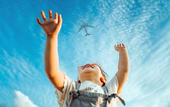 Happy little Asian girl with flower-shaped sunglasses smiling joyfully and raised her hands waving to the aeroplane in the clear blue sky