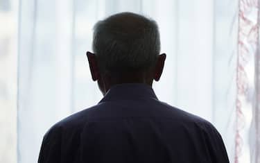 Silhouette of retired man looking through window with transparent curtain standing at home rear view. Loneliness and old human care concept.