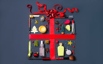 Christmas objects and beauty products laid out in the shape of a gift box with red ribbon on dark gray background. Merry Christmas, Happy New Year, Valentine's Day creative concept of gifts with beauty cosmetic products. Flat lay, copy space.