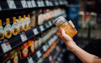 Cropped shot of young woman grocery shopping in supermarket, picking up a bottle of organic honey from the product aisle and reading the nutrition label on the bottle. Routine shopping. Healthy eating lifestyle