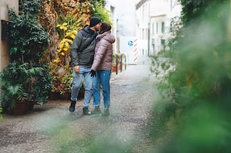 Couple in love kissing on street. In love with warm things, walking along European street with lush greenery, decorated with bright holiday lights. Vacation trips. Merry Christmas and Happy New Year