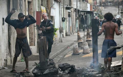 A police officer holds his gun up next to residents on a barricade during the Operacao Contencao (Operation Containment) at the Vila Cruzeiro favela, in the Penha complex, in Rio de Janeiro, Brazil, on October 28, 2025. Hundreds of heavily armed Brazilian police raided slum areas of Rio in a drug raid Tuesday, touching off firefights likened to scenes from a war and leaving at least 64 people dead, Brazilian officials said. (Photo by Mauro PIMENTEL / AFP)
