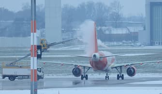 SCHOENEFELD, GERMANY - NOVEMBER 28: In this view through a pane of glass, a de-icing vehicle de-ices a passenger plane as snow lies on the tarmac at Berlin International Airport (BER) on November 28, 2023 in Schoenefeld, Germany. A nationwide snowfall led to disruption in some regions. (Photo by Sean Gallup/Getty Images)