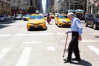 An elderly man crosses the street in Manhattan in front of iconic yellow taxi cabs, having arrived in America for the first time since he came as a member of the Royal Navy during World War 2.  The gentleman is dressed in a blue shirt and navy trousers and carries a walking stick to steady him as he makes his way over the pedestrian crossing on a typical New York Street on a sunny day in Summer.
