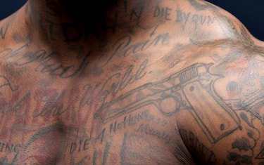 African American man's shoulder and chest view with tattoos of gun, skull, and different phrases.