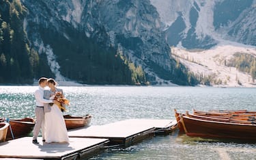 The bride and groom walk on a wooden boat dock at Lago di Braies in Italy. Wedding in Europe, at Braies lake. Newlyweds walk, kiss, cuddle against the backdrop of rocky mountains.