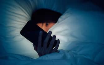 Woman hiding under the blanket, chatting and surfing the internet with smart phone at late night on bed. Working late concept. Reading work email at late night. Busy city life. Working late at night. Online bullying and hate speech concept. Bad news.