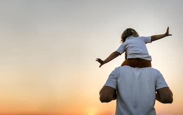 Dad carries baby toddler on his shoulders at sunset in field