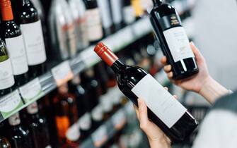 Over the shoulder view of woman walking through liquor aisle and choosing bottles of red wine from the shelf in a supermarket