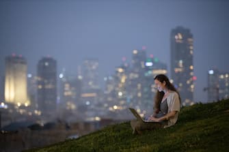 Smiling woman working remotely on laptop in a public park with high-rise buildings seen in background at dusk time.