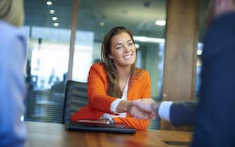 a young graduate sits across the table from her interview panel  full of confidence and positivity energy . She is holding her cv and smiling at the interview panel before her and shakes hands with the new boss.She is wearing blue trousers with an orange suit jacket . in the foreground we can see the back of two of the panel , a man and a woman.